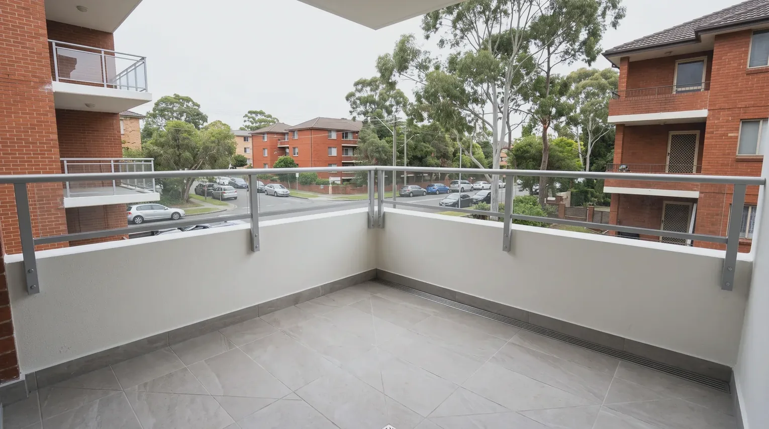 A newly completed apartment balcony with new stone-look tiles, a frameless glass balustrade and a potted olive tree, overlooking leafy Sydney rooftops at golden hour