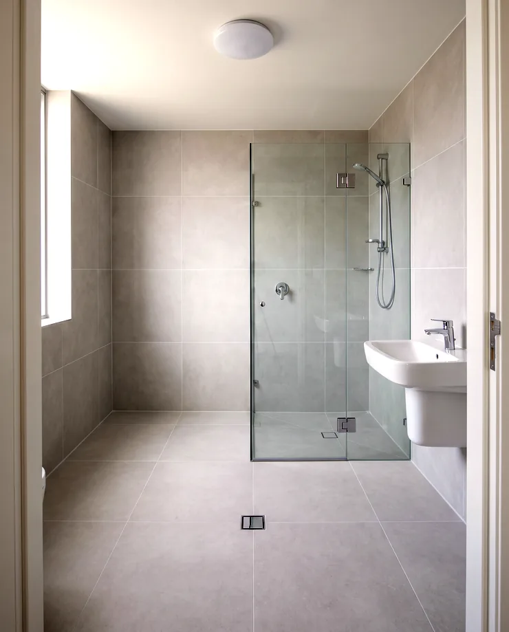 A finished apartment bathroom after re-waterproofing, with pale grey tiling, a chrome shower set and a clean silicone line at the floor junction
