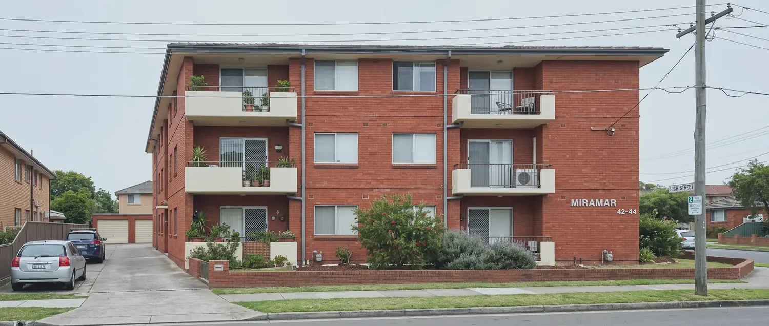 A three storey red brick apartment building on an ordinary Sydney street, photographed from the footpath opposite on an overcast day